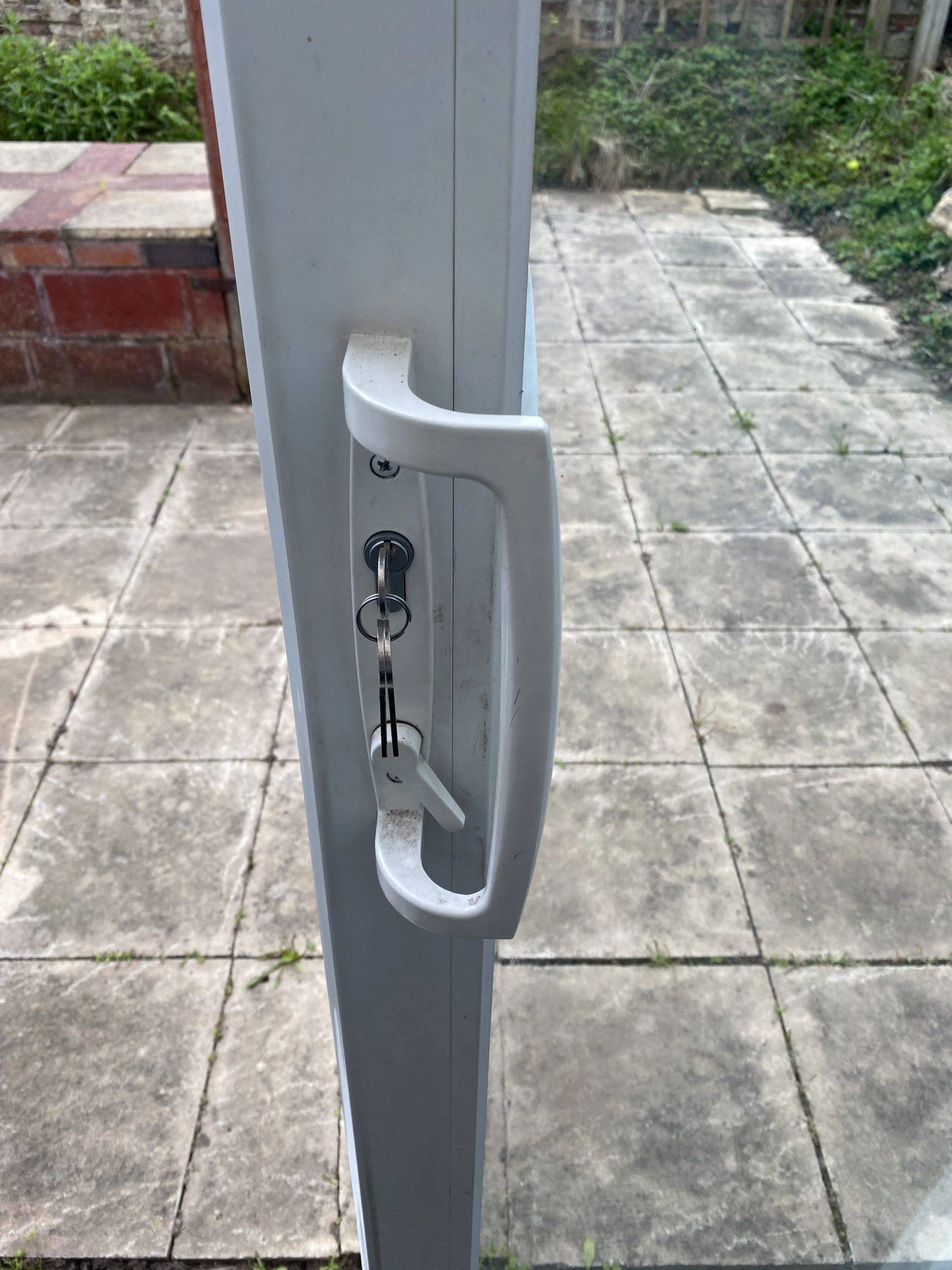 White patio door handle with key inserted, opening onto a paved garden area fixed by an Emergency Locksmith